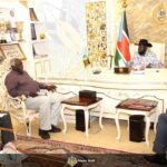 A wide-shot photograph shows South Sudan's President Salva Kiir Mayardit, wearing his signature black Stetson hat and a patterned shirt, seated behind a large, ornate white and gold desk in an office. He is meeting with three officials: two men are seated in decorative white armchairs to the left, and one man is seated to the right. The room is formal, featuring the national flag of South Sudan, the Presidential seal on the wall, and shelves filled with trophies and framed photos. A "Press Unit" watermark is visible across the bottom of the image.