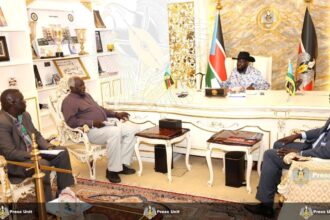 A wide-shot photograph shows South Sudan's President Salva Kiir Mayardit, wearing his signature black Stetson hat and a patterned shirt, seated behind a large, ornate white and gold desk in an office. He is meeting with three officials: two men are seated in decorative white armchairs to the left, and one man is seated to the right. The room is formal, featuring the national flag of South Sudan, the Presidential seal on the wall, and shelves filled with trophies and framed photos. A "Press Unit" watermark is visible across the bottom of the image.