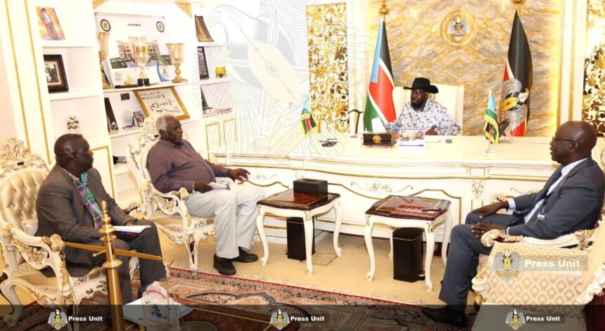 A wide-shot photograph shows South Sudan's President Salva Kiir Mayardit, wearing his signature black Stetson hat and a patterned shirt, seated behind a large, ornate white and gold desk in an office. He is meeting with three officials: two men are seated in decorative white armchairs to the left, and one man is seated to the right. The room is formal, featuring the national flag of South Sudan, the Presidential seal on the wall, and shelves filled with trophies and framed photos. A "Press Unit" watermark is visible across the bottom of the image.
