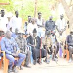 A large group of men and women gathered outdoors for a formal meeting, seated in orange and blue chairs under a tree with traditional staffs.